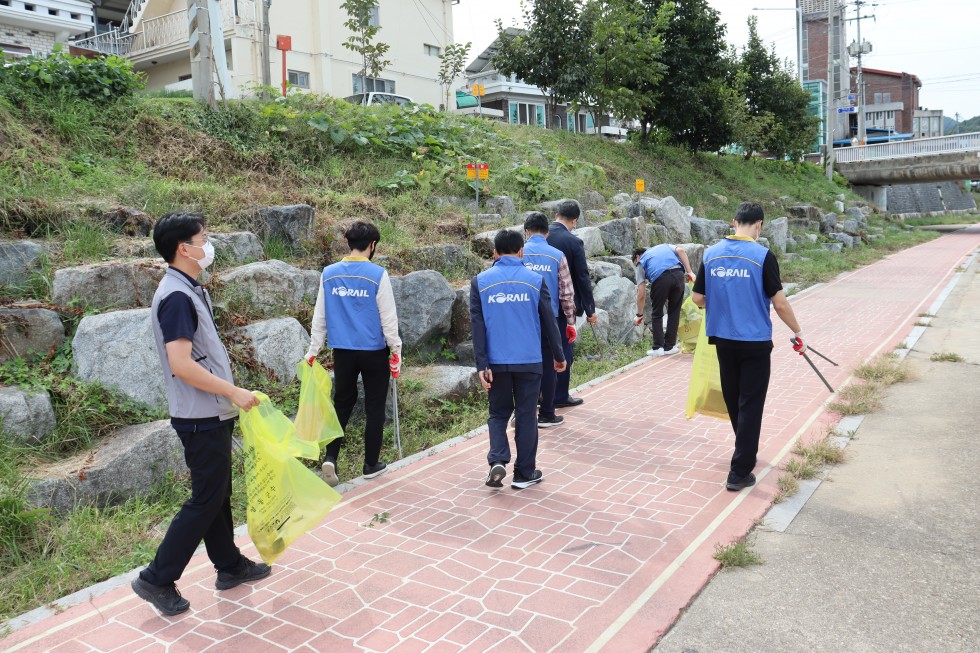 코레일 직원과 복지관 직원이 함께 영동군 천변을 걸으면서 쓰레기를 집게로 집어 쓰레기 봉투에 넣으면서 환경미화 작업을 진행하고 있는 모습입니다.